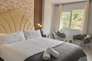 A hotel room with a large bed, two armchairs, a small table, and a window with a view of greenery. - Marbella Coastal Comfort (Marbella)