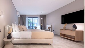 A modern bedroom with a large bed, a TV mounted on the wall, and a dining area visible through the window.