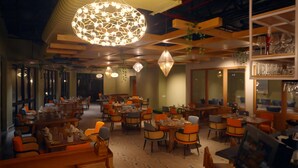 A restaurant with multiple tables and chairs, hanging light fixtures, and a bar area with shelves of glasses. - The Solitaire bliss (Tinsukia)