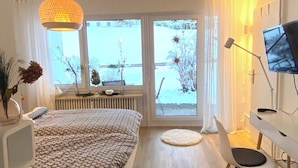 Ein Schlafzimmer mit Bett, Fernseher, Fenster und Blick auf Bäume sowie einer Heizung.