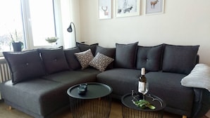 Ein modernes Wohnzimmer mit einem grauen Ecksofa, gemusterten Kissen, einem runden Couchtisch und einer Weinflasche.
