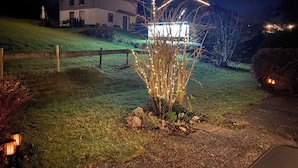 Ein Haus mit einer gut beleuchteten Einfahrt bei Nacht, einem Holzzaun und einer sanften, grasbewachsenen Böschung.