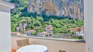 A balcony with wicker furniture and a table, overlooking a village with buildings and a cliff. - Appartamento a Amalfi ID 3059 (Amalfi)