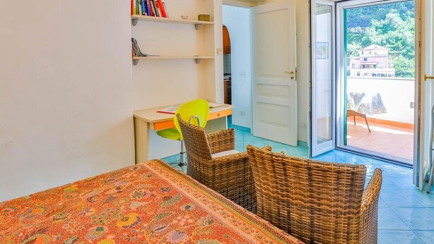 Una habitación con una mesa de comedor, sillas de mimbre, una estantería y una puerta que da a un espacio exterior con vistas a edificios y árboles.