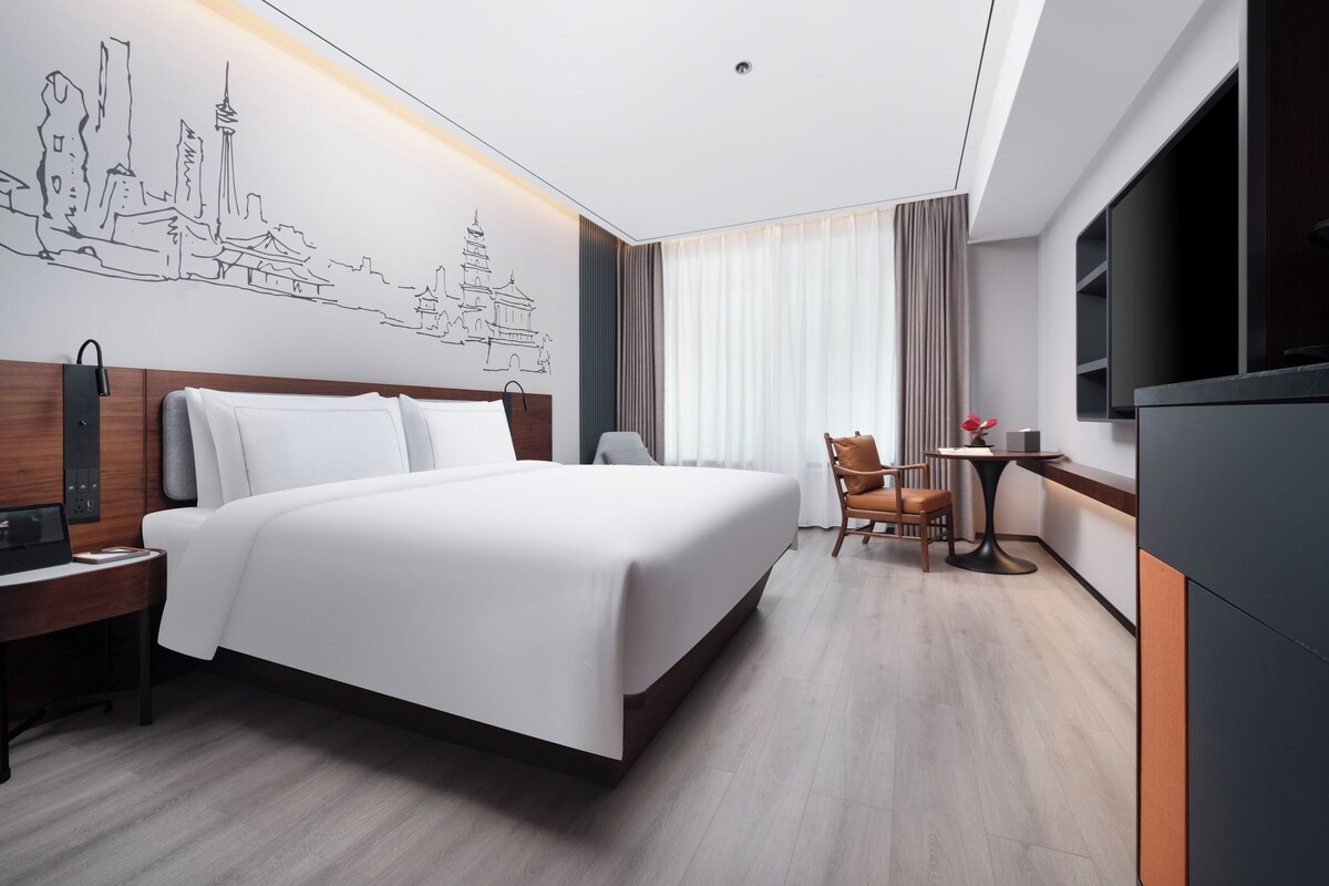 Une chambre d’hôtel moderne dotée d’un grand lit, d’un bureau, d’une chaise, d’une télévision et d’une œuvre d’art murale représentant un paysage urbain.