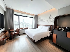 A hotel room with a bed, a desk, a chair, a window with a city view, and a minibar. - UrCove by Hyatt Xi'an Great Tang All Day Mall (Xi'an)