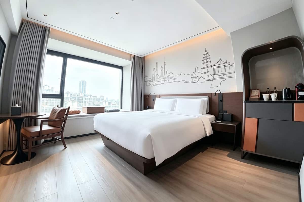 A modern hotel room with a large bed, a desk, a chair, a nightstand, and a view of the cityscape.