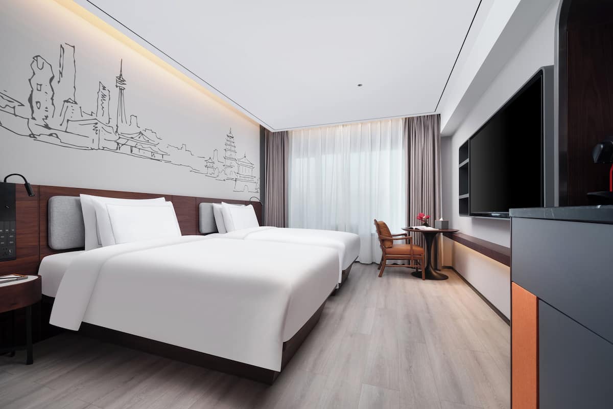 A modern hotel room with a large bed, a desk, a chair, a TV, and a wall art depicting a cityscape.