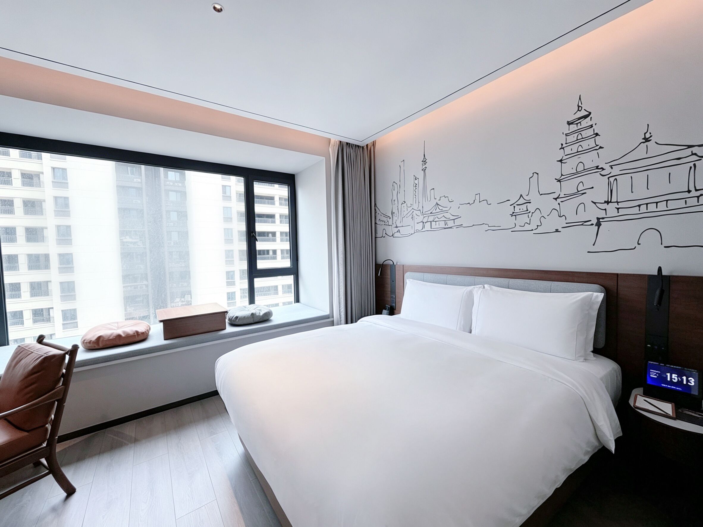 A hotel room with a large bed, a chair, a window with a city view, and a wall with a cityscape mural.