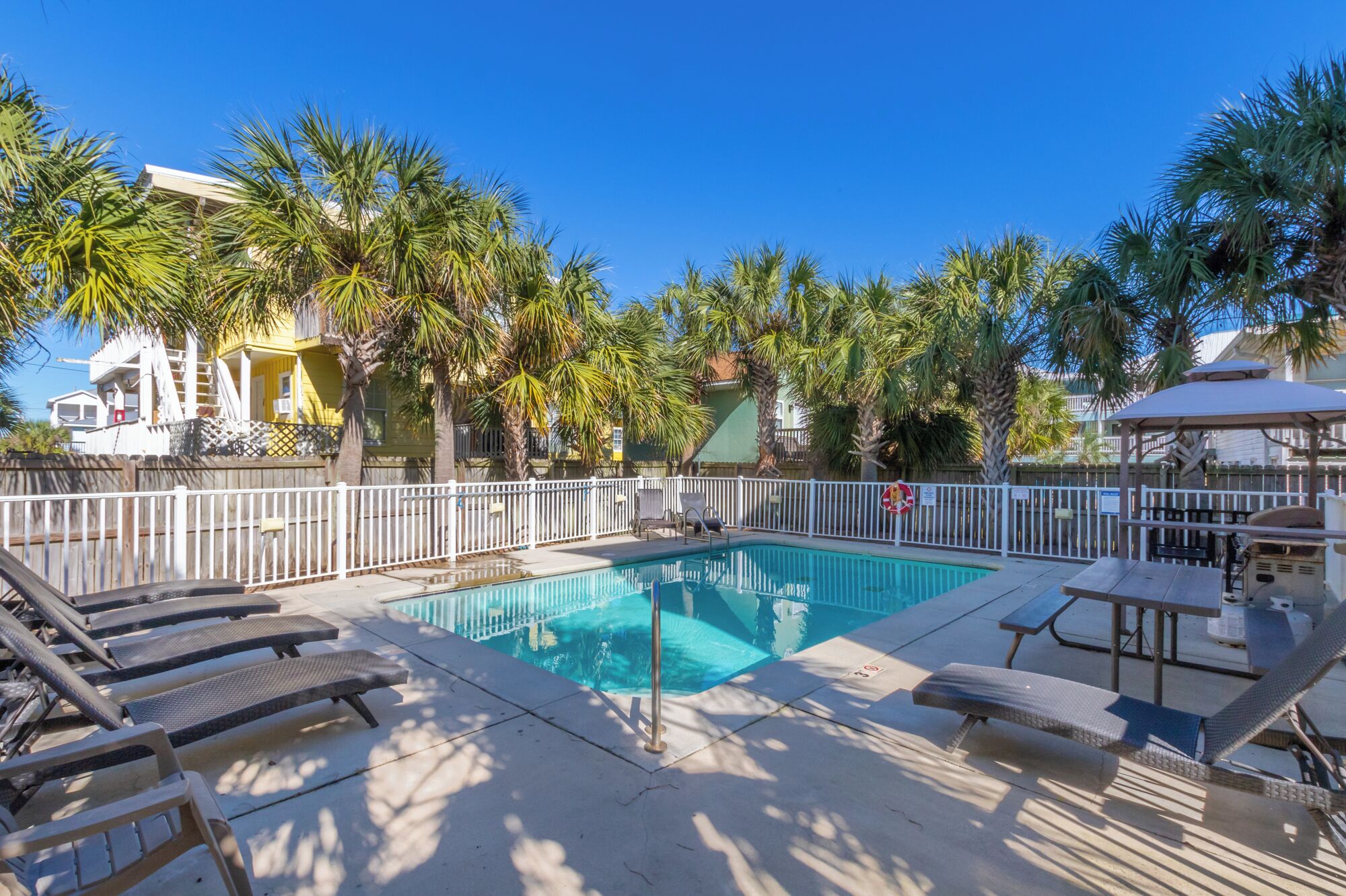 A swimming pool with lounge chairs and a fenced area, surrounded by palm trees and a building with a yellow facade.