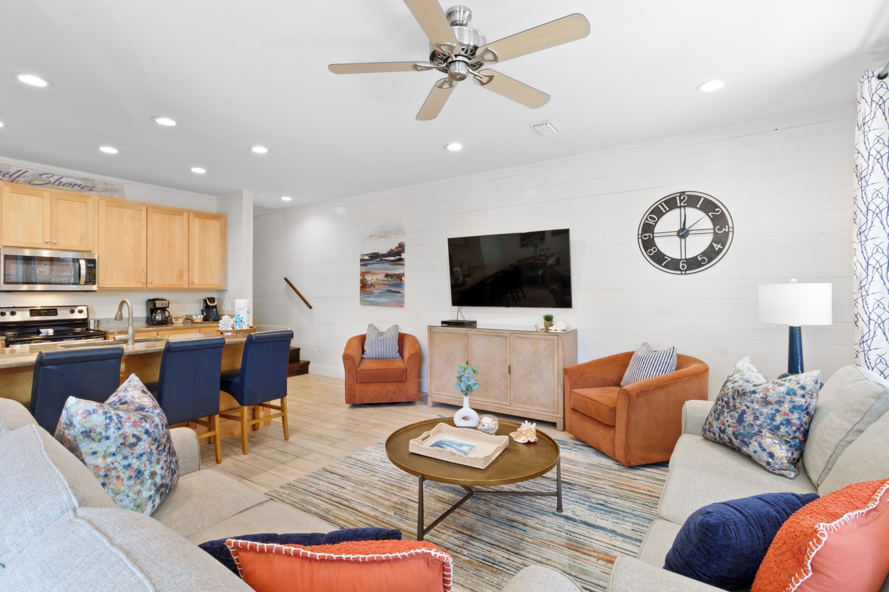A modern living room with a sofa, coffee table, and chairs. There is a kitchen area with a microwave and stove. A ceiling fan and clock are visible.