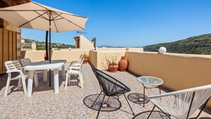 A terrace with a table and chairs, a large umbrella, and potted plants. - Countryside Apartment 2 With Shared Swimming Pool (Kissamos)