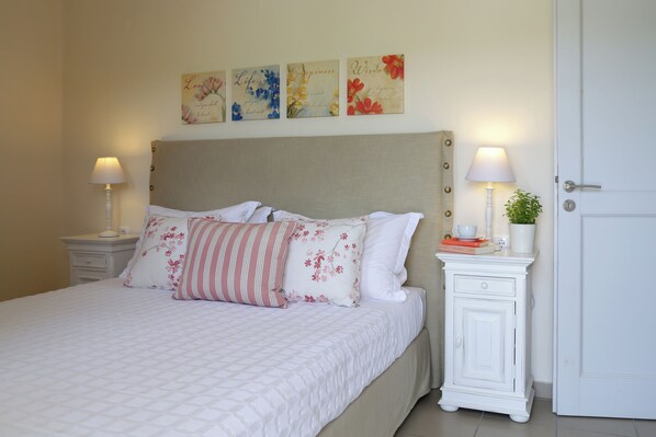 A neatly made bed with floral pillows, a bedside table with a lamp, and a small cabinet with a plant. - Skopelos Summer Homes (Skopelos)