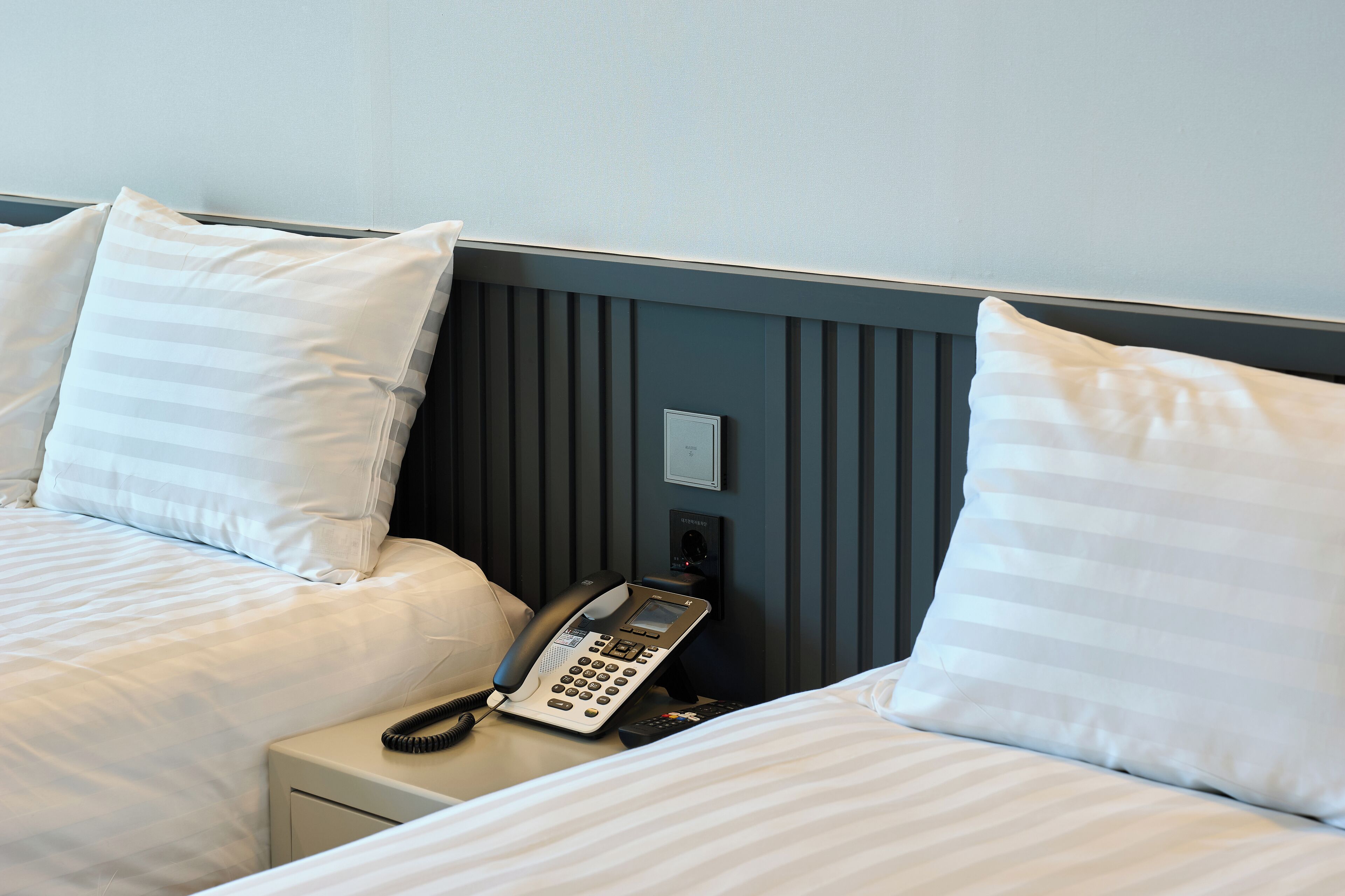 A hotel room with two beds, each with white striped bedding and pillows. There is a bedside phone on the nightstand between the beds.