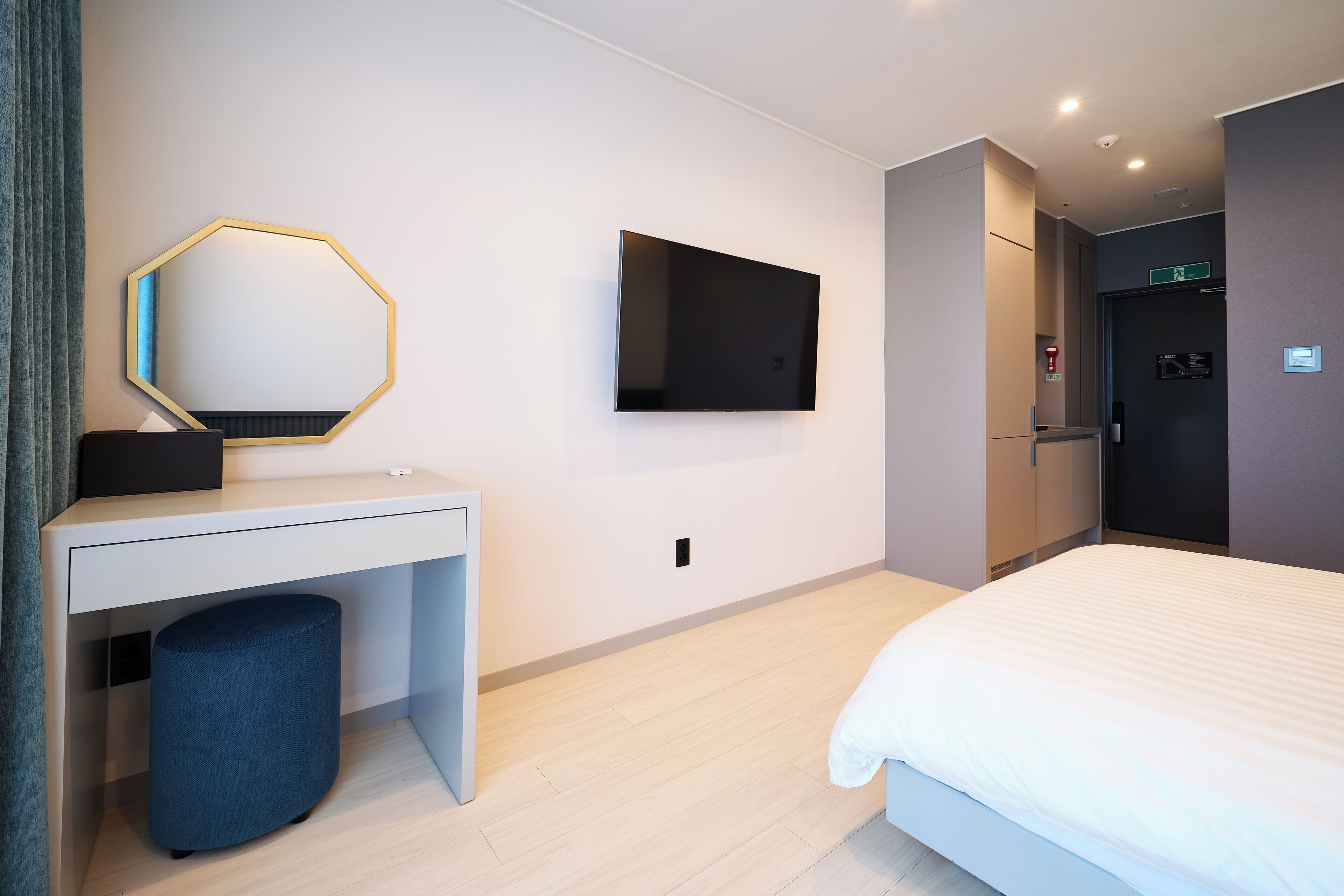 A modern hotel room with a bed, a desk with a mirror, a flat-screen TV, and a door with an exit sign.