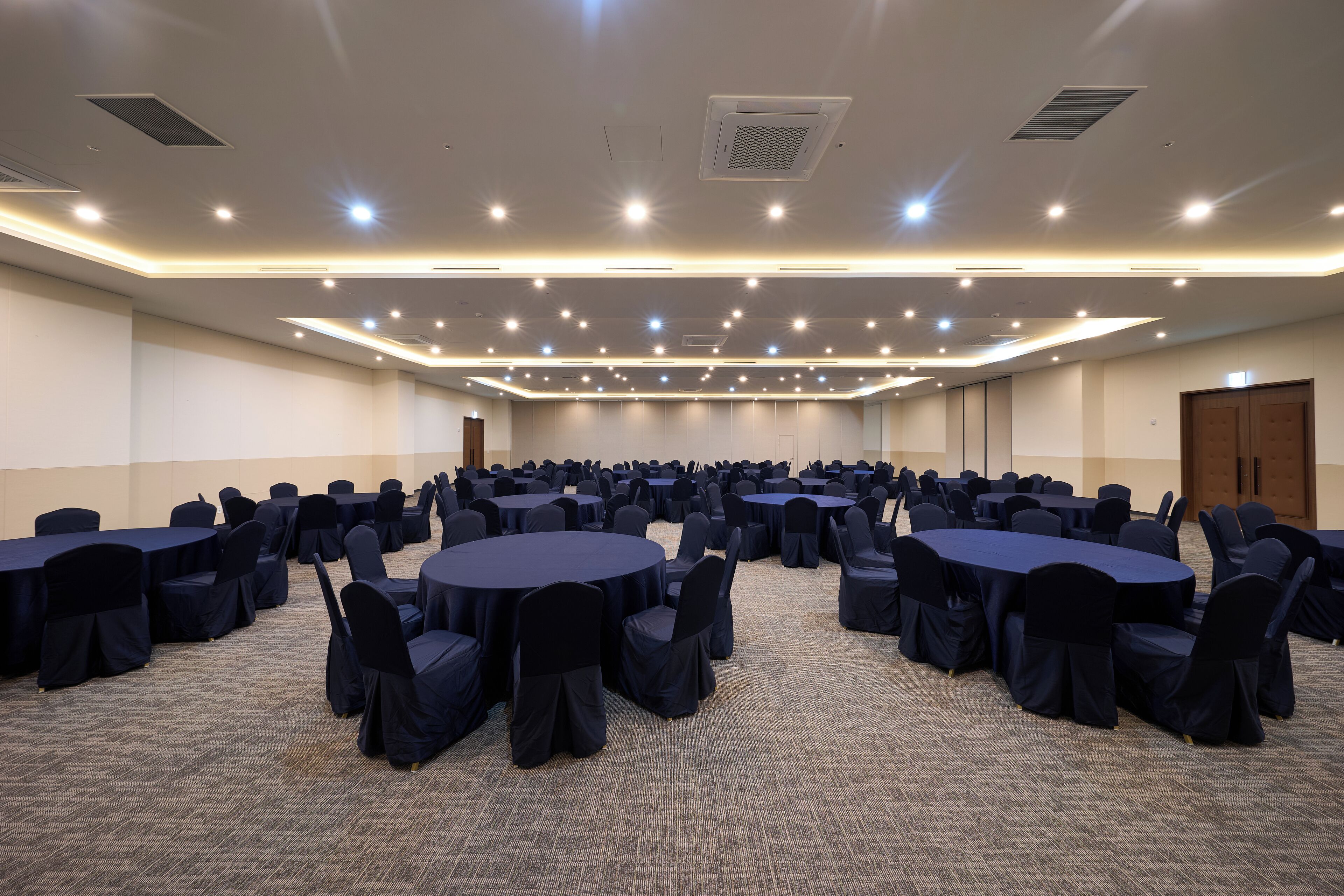 A conference room with multiple round tables and chairs, carpeted floor, and recessed lighting.