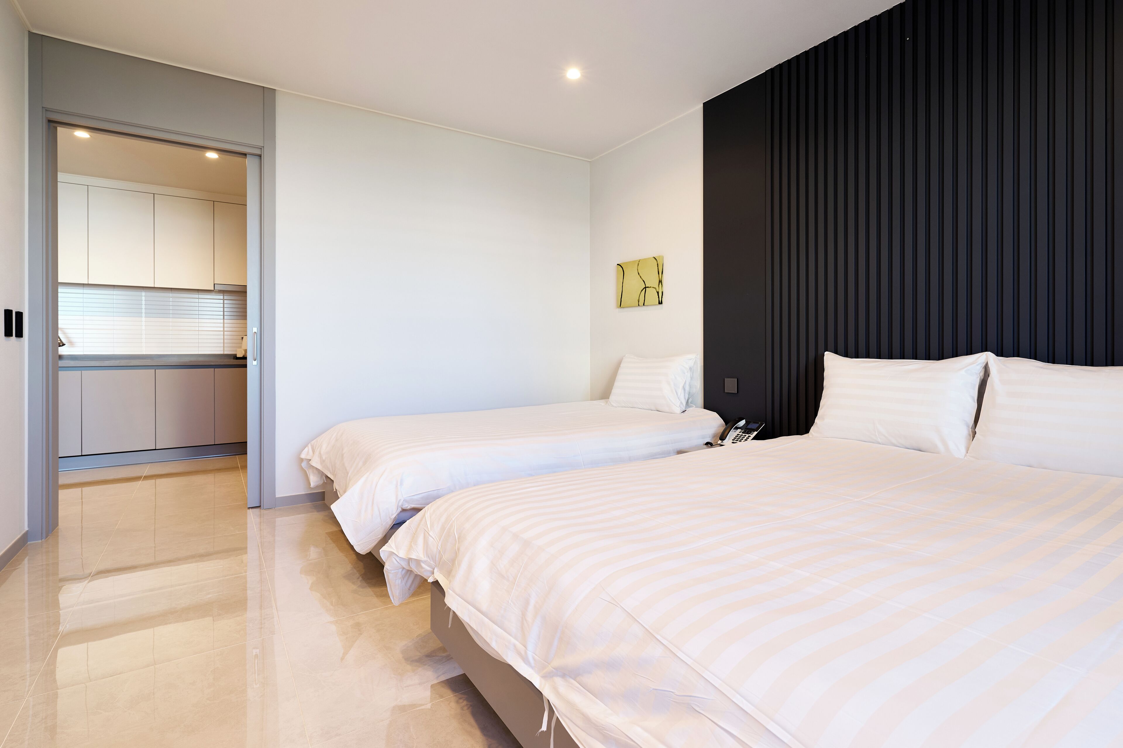 Et moderne hotelværelse med to senge, en mørk væg og et køkkenområde, der kan ses gennem en åben dør.