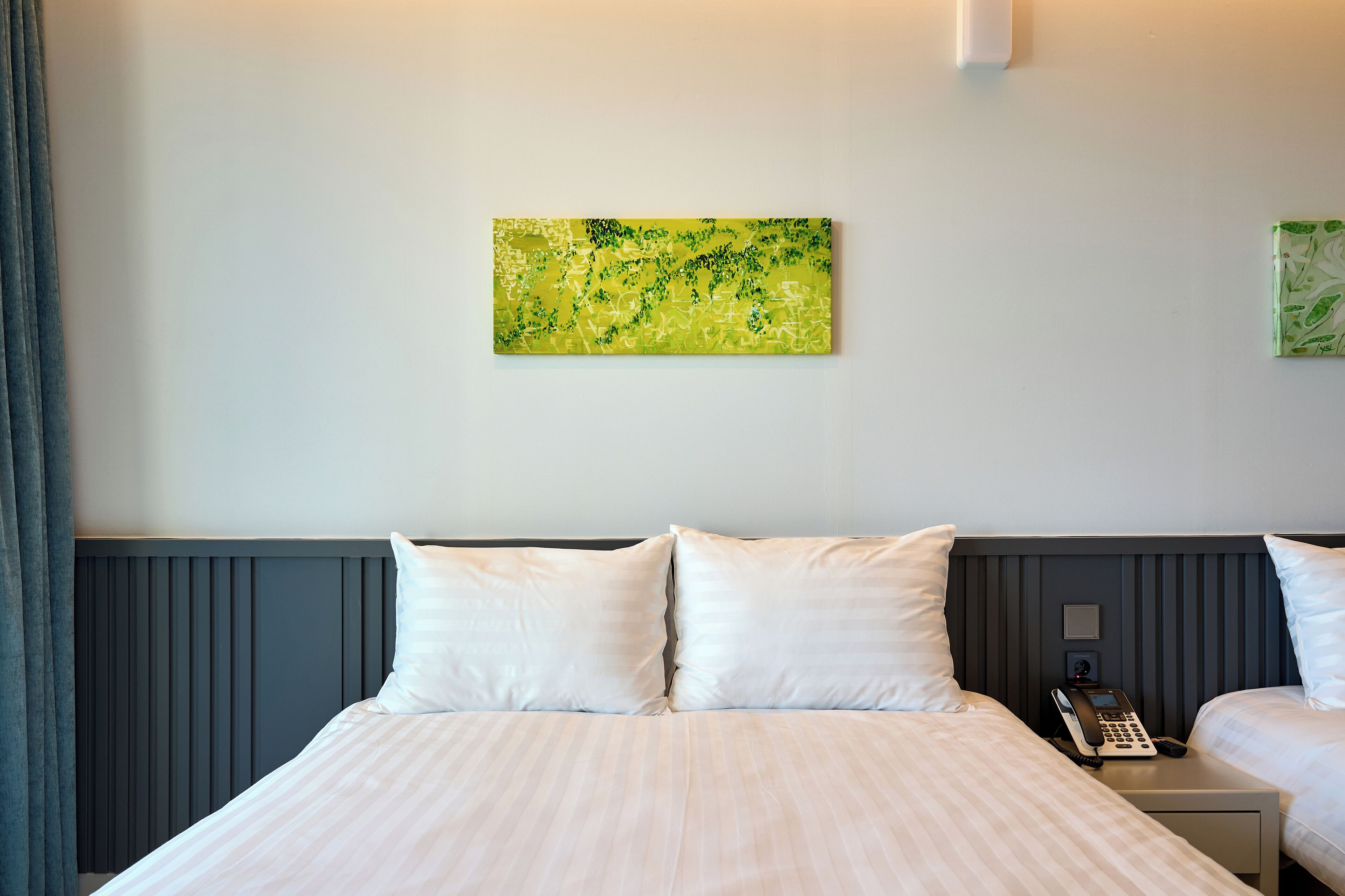 Hotellihuone, jossa on suuri sänky, kaksi tyynyä, yöpöytä, jossa on puhelin, ja taulu seinällä.