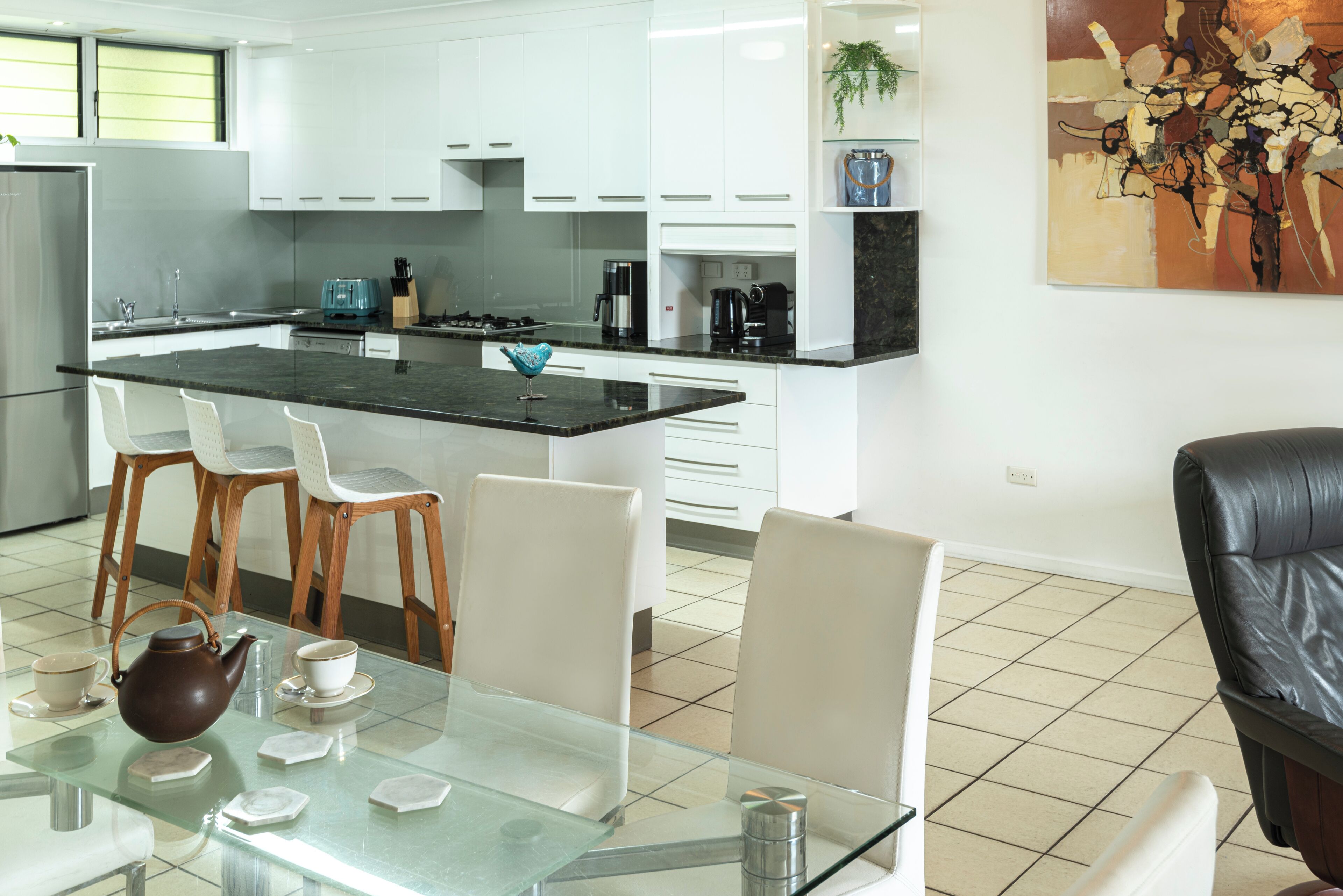 Een moderne keuken met witte kasten, een zwart aanrecht en een glazen eettafel met stoelen.
