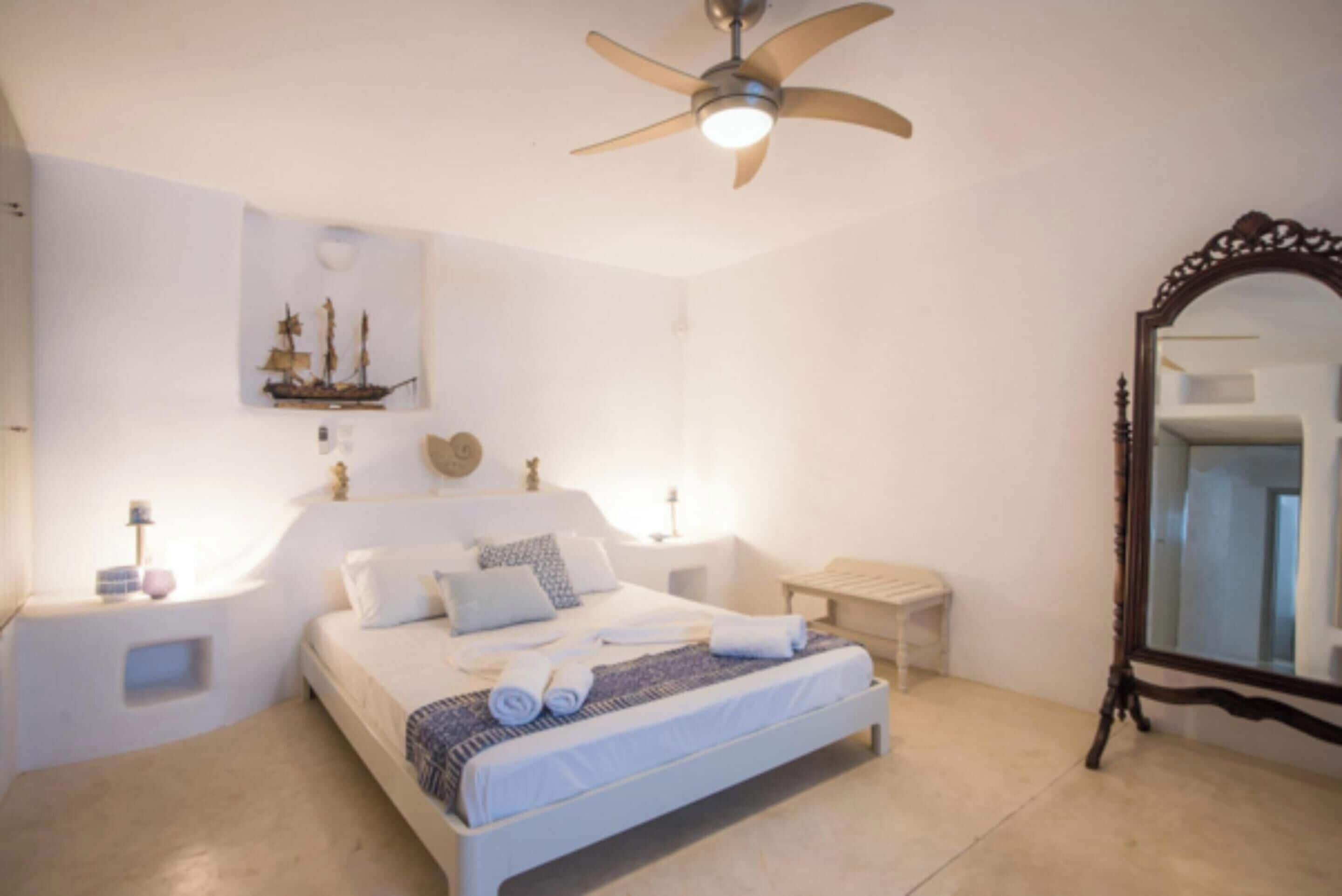 A bedroom with a bed, a ceiling fan, a nightstand, a bedside table, a mirror, and a wall decoration.