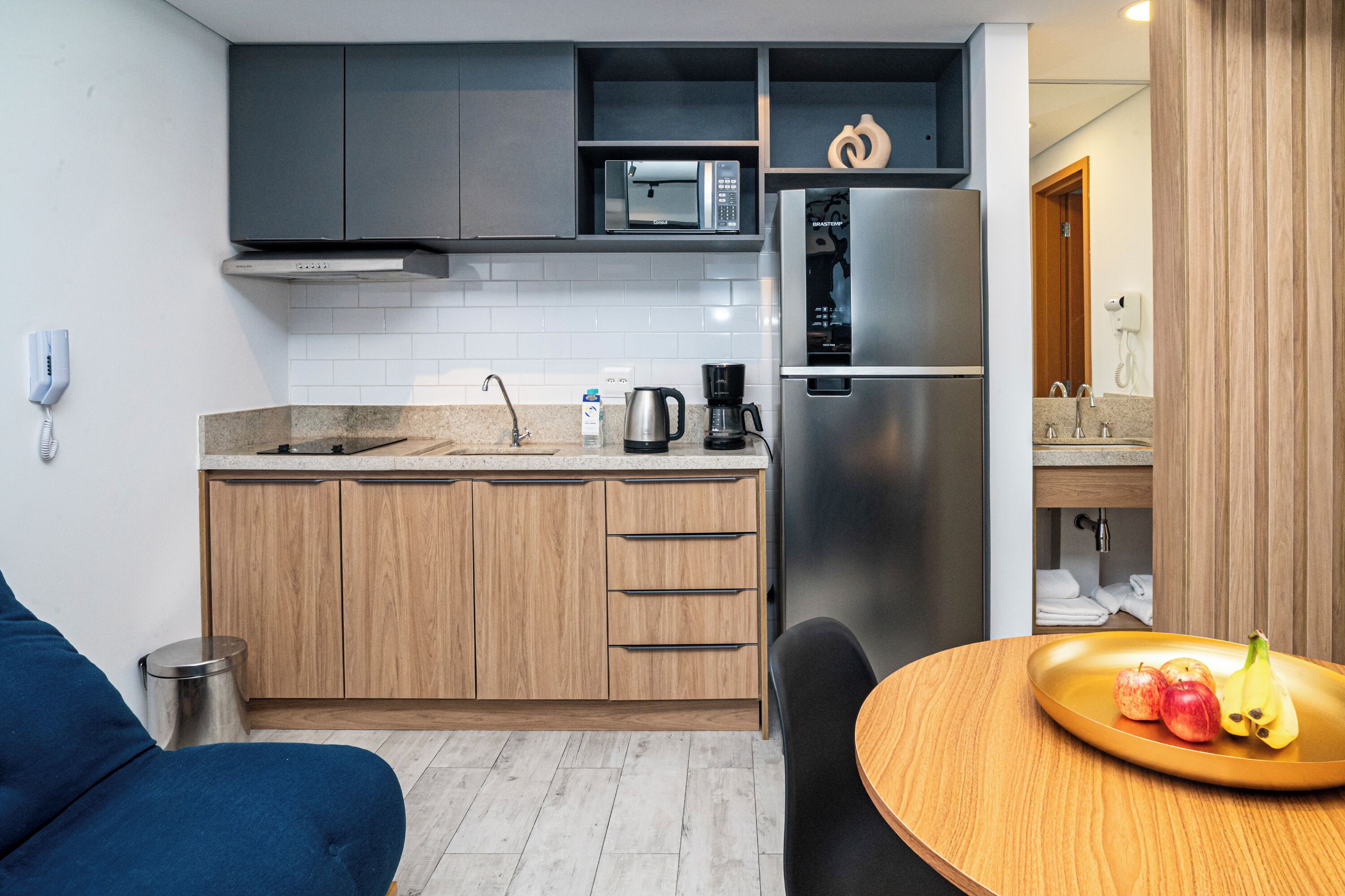 Cocina compacta con microondas, refrigerador y cafetera. Se aprecia un mueble de madera y un pequeño bote de basura. Se ve una mesa de comedor con un plato de fruta.