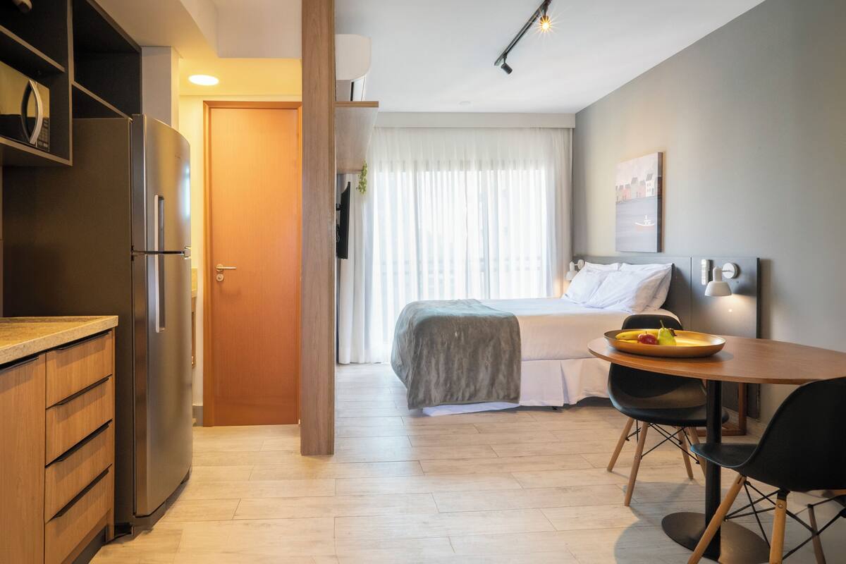 A modern hotel room with a kitchenette, a bed with a gray blanket, a dining table with chairs, and a cityscape painting on the wall.