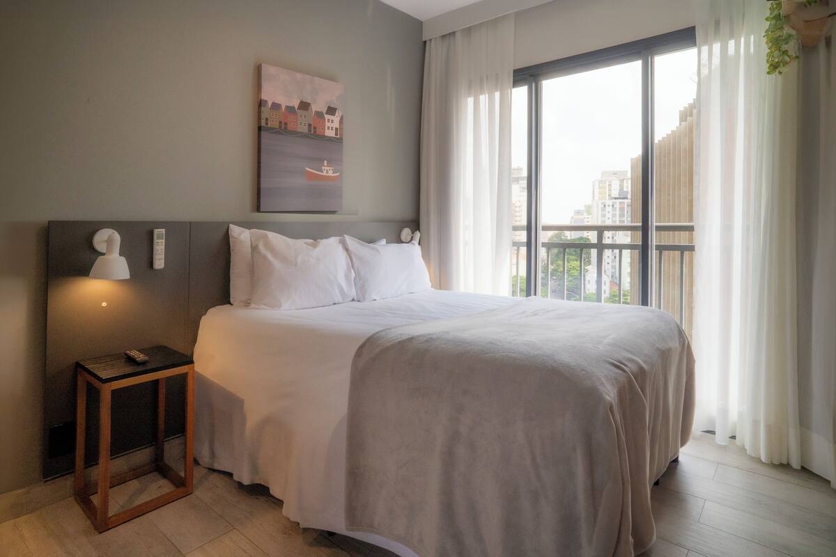 A neatly made bed with white linens, a bedside table with a remote control, and a painting on the wall.