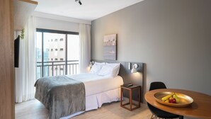 A hotel room with a bed, a dining table with fruit, a small side table, and a view of buildings through the window.