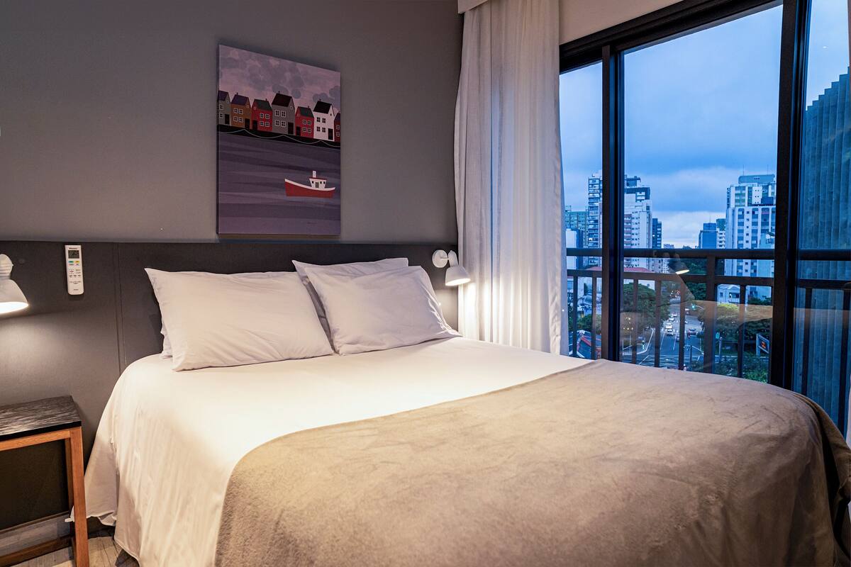 A hotel room with a large bed, a nightstand, a wall-mounted lamp, and a window with a city view.