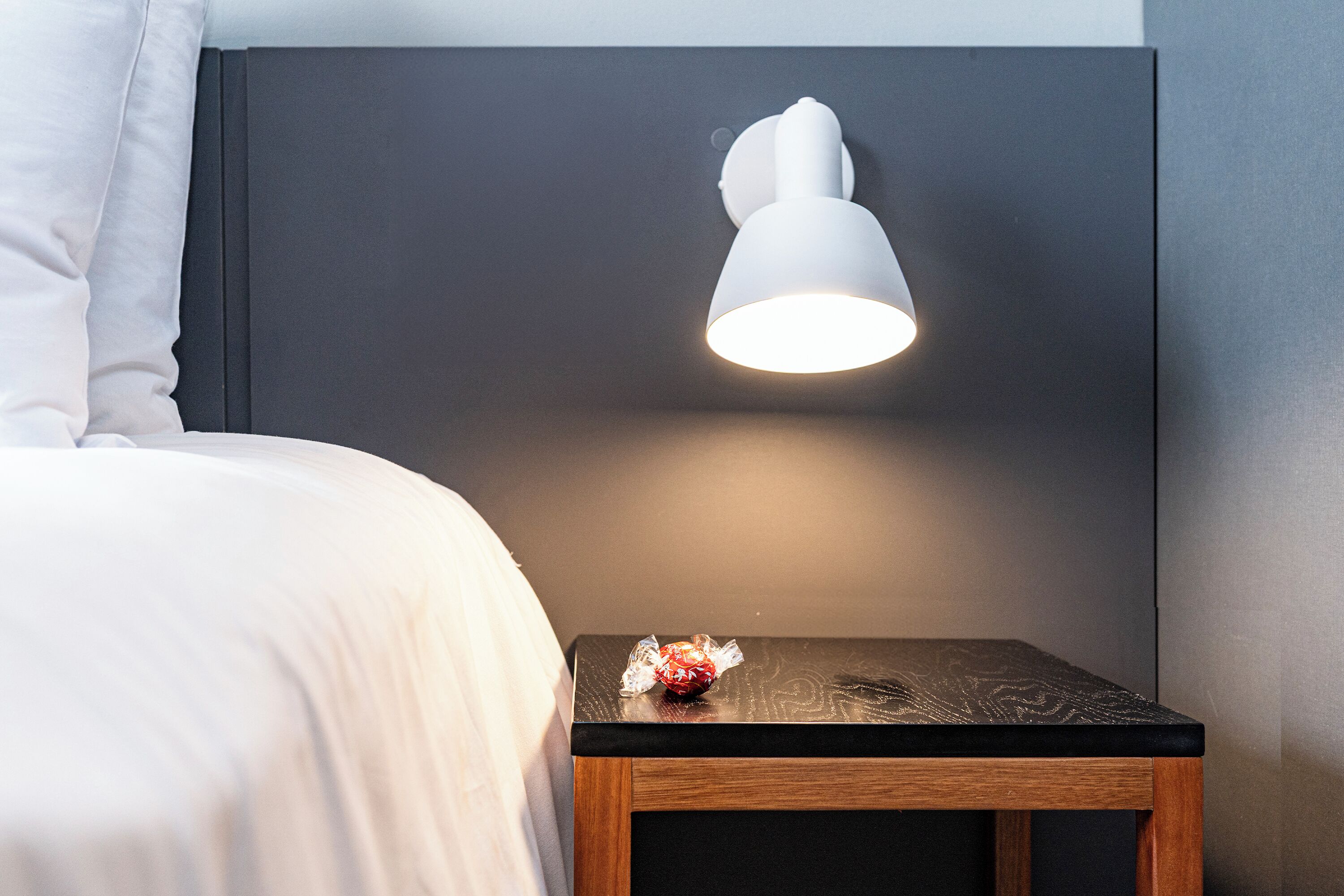 Un lit bien fait, avec du linge de lit blanc, une petite table de chevet en bois foncé et une lampe blanche fixée au mur.