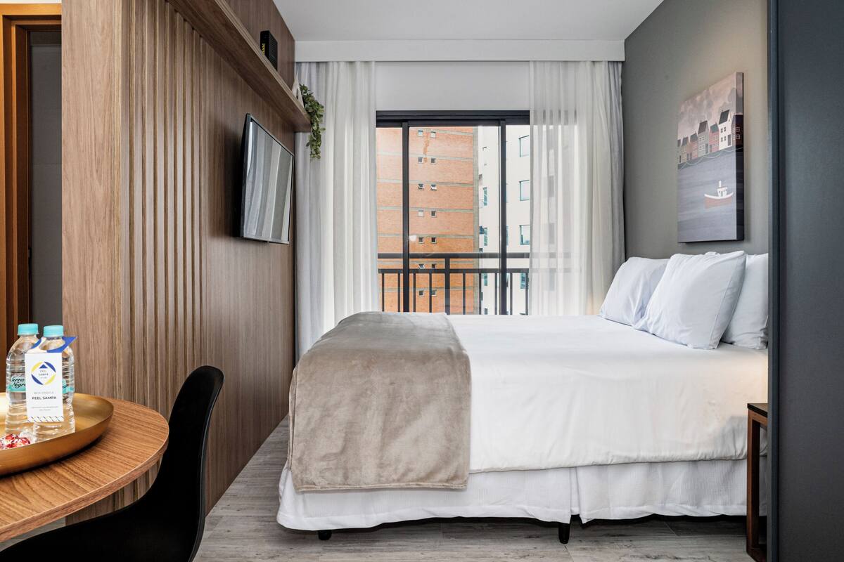 A hotel room with a bed, a TV mounted on the wall, a window with a view of buildings, a wooden panel wall, and a small table with water bottles.