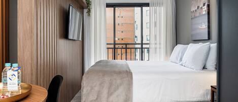 A hotel room with a bed, a desk with a chair, a TV mounted on the wall, and a window with a view of buildings.
