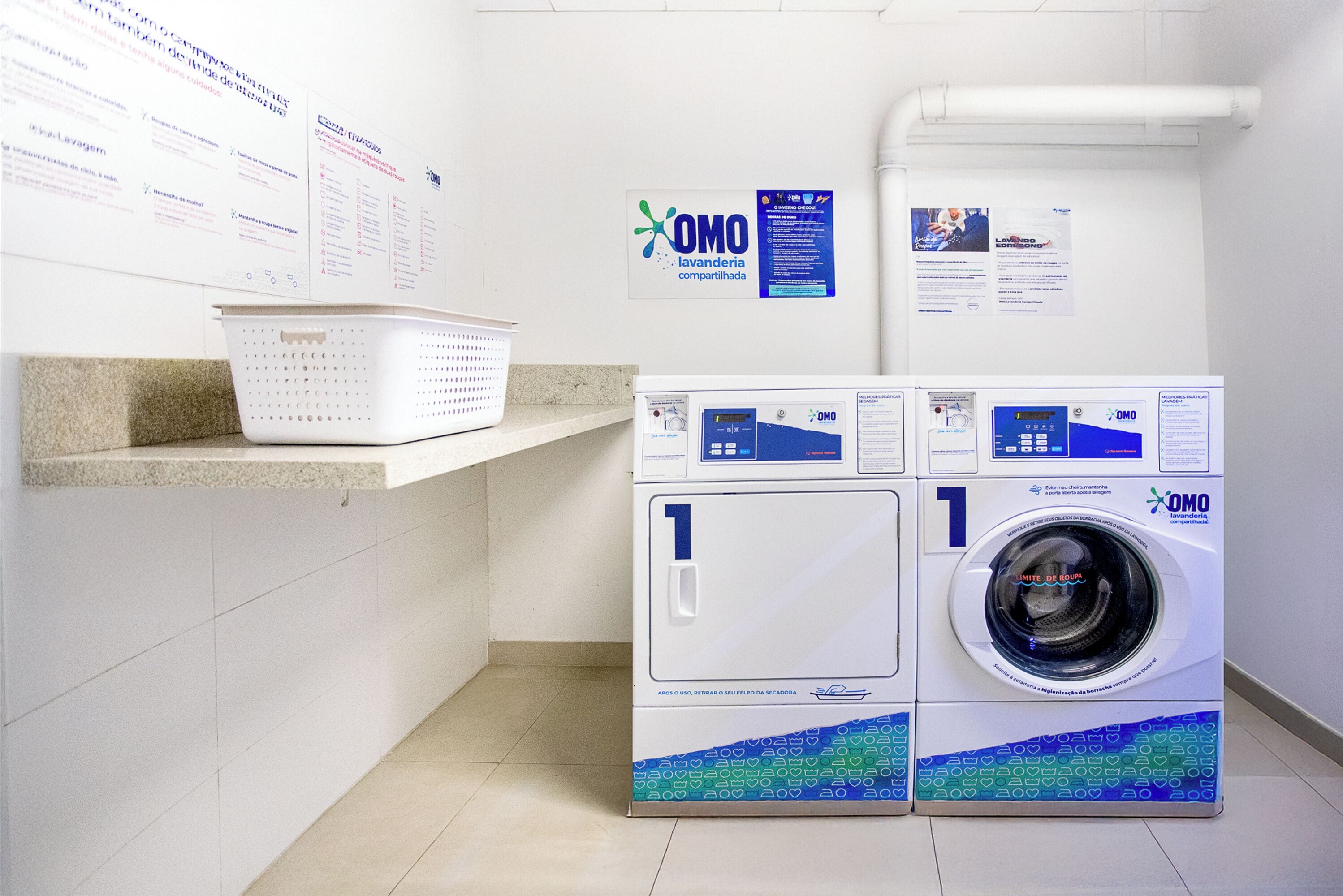 A laundry area with two OMO branded washing machines, a white wall with informational posters, and a white tiled floor.