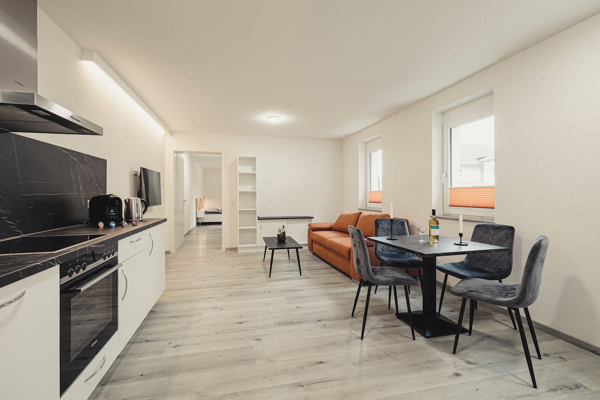 A modern kitchen with a black countertop, built-in oven, and a stainless steel range hood. There is a dining area with a table and chairs, and a sofa in the background.