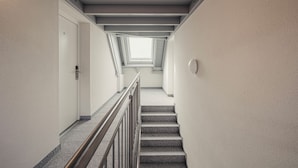 Egy keskeny, jól megvilágított lépcsőház, fém korláttal, amely egy olyan platformhoz vezet, ahol egy tetőablak található.