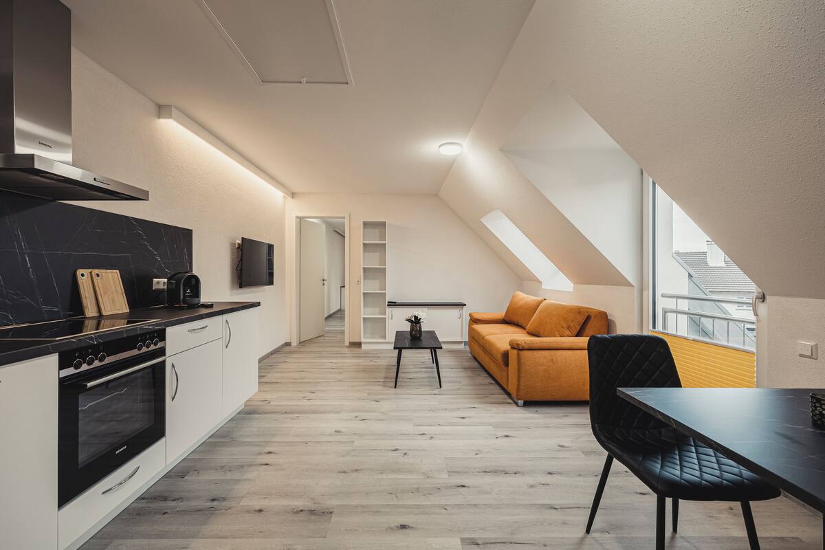 A modern kitchen with a black countertop, white cabinets, and a built-in oven. There is a dining area with a black table and a yellow sofa.