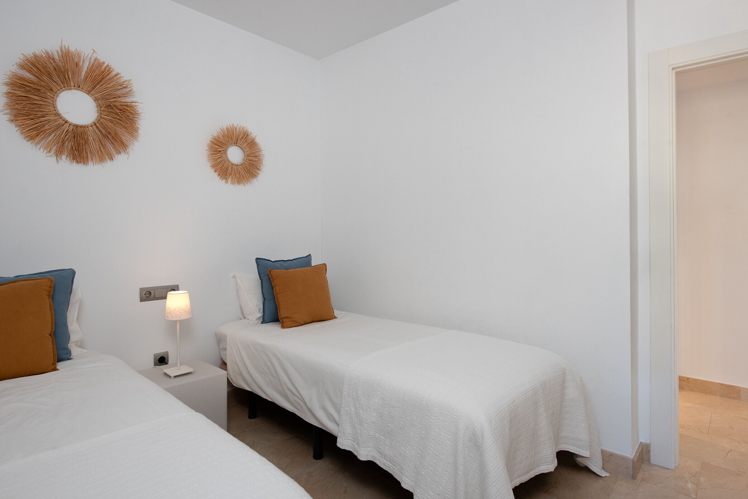 Habitación de hotel con dos camas individuales, una mesita de noche con lámpara y dos adornos colgantes en la pared.