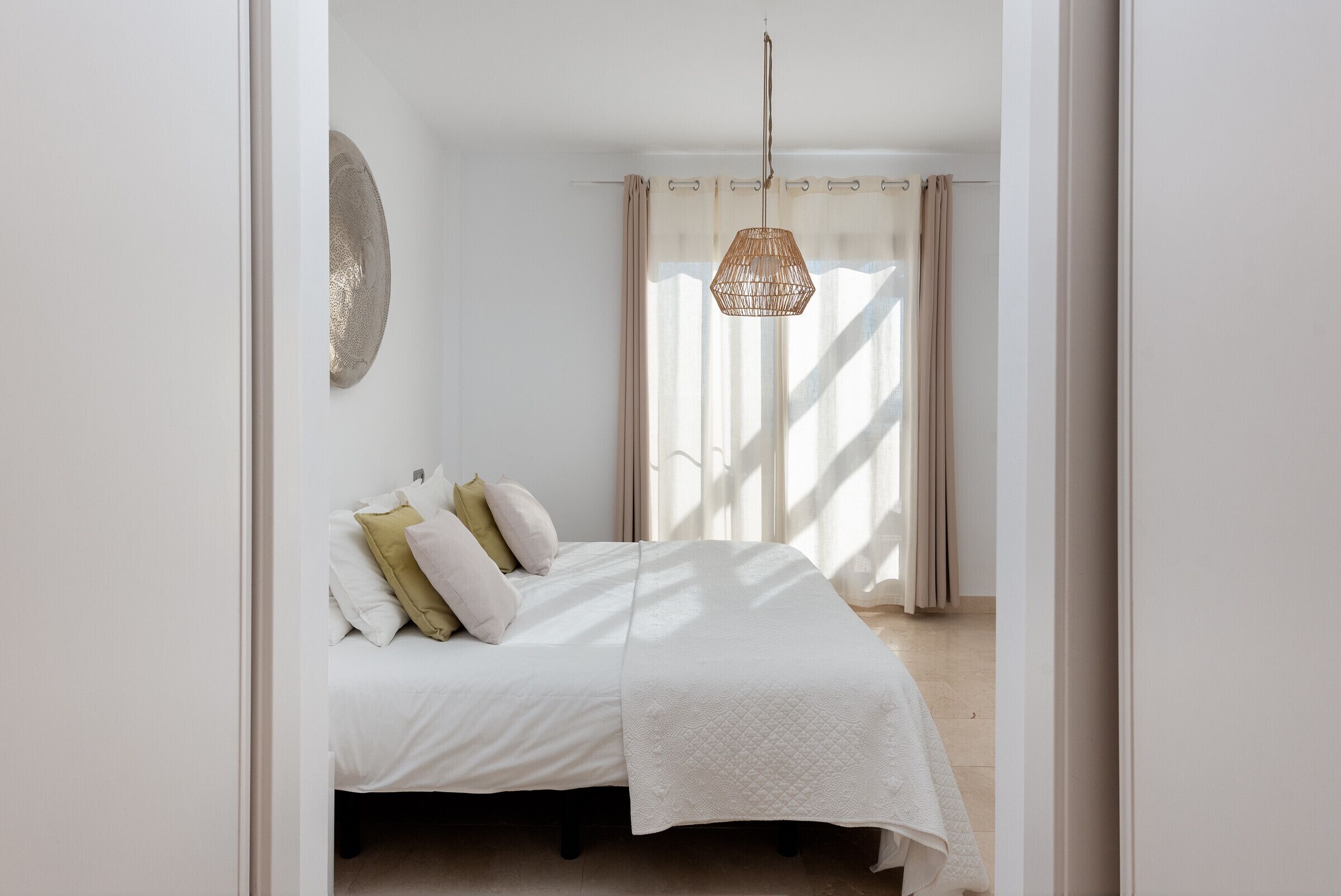 A bedroom with a bed, pillows, a hanging light fixture, and a view of a window with curtains.