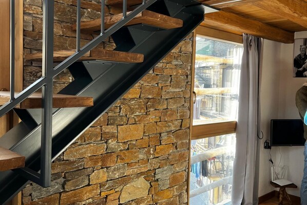 Una escalera moderna con pared de piedra y vigas de madera, que conduce a una habitación con un ventanal grande y una televisión de pantalla plana.
