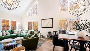 A spacious living room with a green sectional sofa, a dining table with chairs, and large windows offering a view of autumn trees. - Pet-Friendly Modern Retreat · Woods · Ellison Bay (Ellison Bay)
