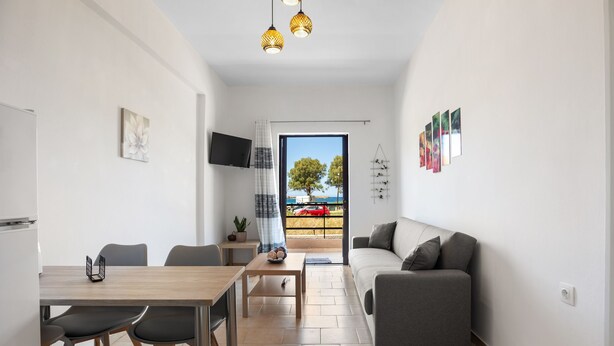 A modern living room with a grey sofa, a wooden dining table with chairs, a TV mounted on the wall, and a balcony with a view of the sea. - Sun Beach Apartment in Chania 3 (Chania)