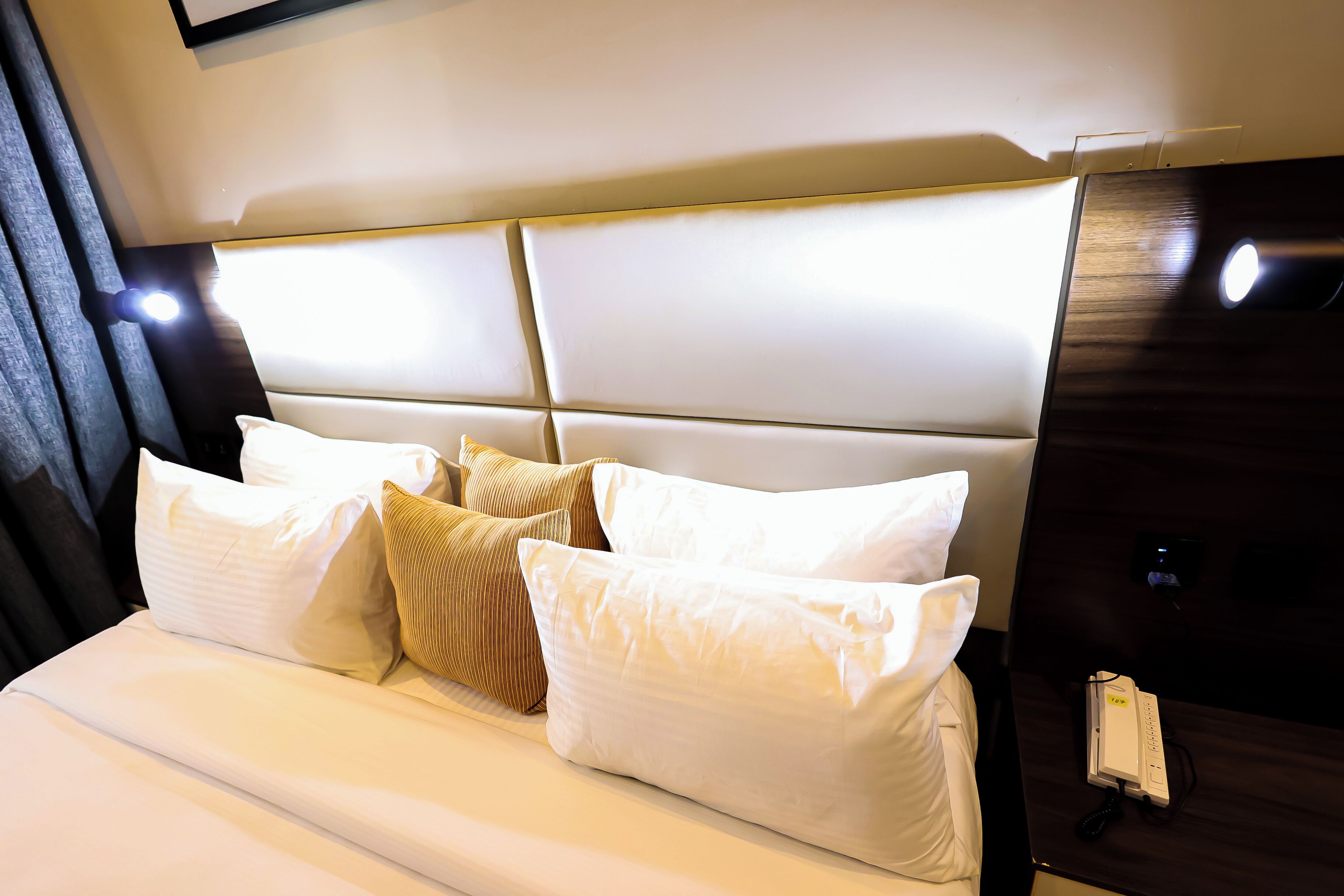 Una habitación de hotel con una cama grande, varios almohadones, una mesita de noche con teléfono, y una lámpara de pared.