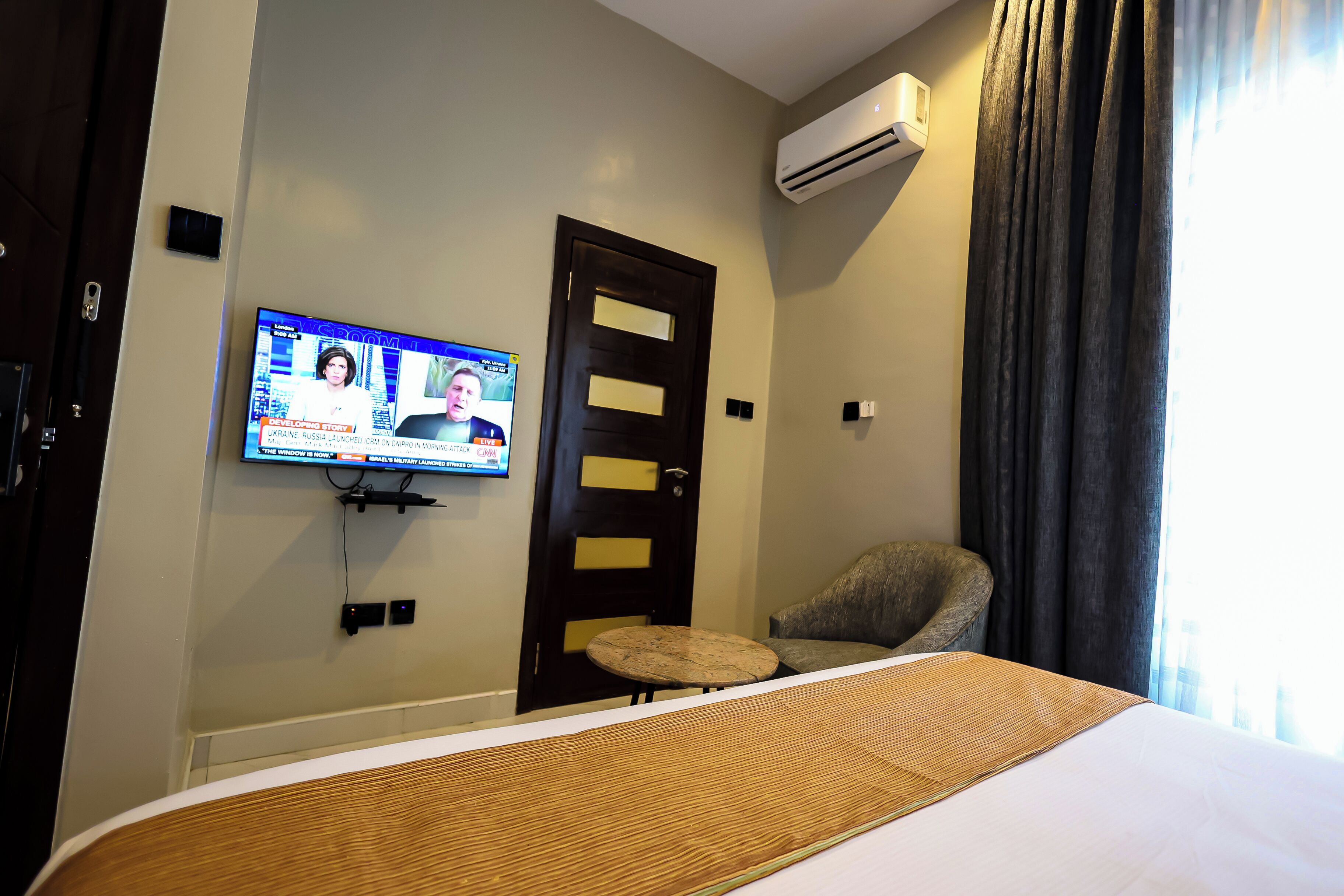 Une chambre d’hôtel avec un lit, un téléviseur fixé au mur, une porte et une chaise.