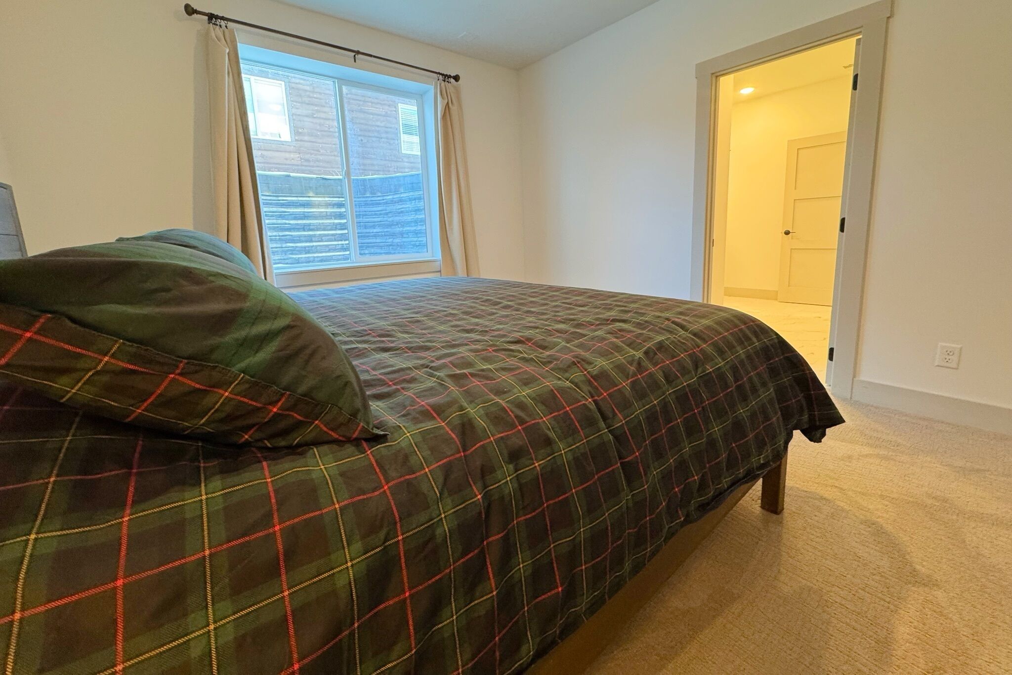 Une chambre à coucher avec un lit, une fenêtre avec des rideaux, un sol recouvert de moquette et une porte donnant sur une autre pièce.