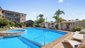 Una piscina con sillas de descanso y una zona de estar cubierta, rodeada de edificios y palmeras.