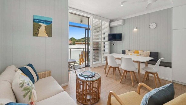 Una sala de estar moderna con un sofá, una mesa de comedor y sillas. Hay un televisor montado en la pared y una ventana con persianas.