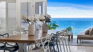 A dining area with a wooden table and white chairs, two white vases with flowers, and a view of the ocean through a glass door. - Epicview - Incredible views, Pets stay free (SAPPHIRE BEACH)