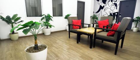 A room with a wooden floor, white walls, two windows with curtains, a table with chairs, and several potted plants.