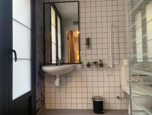 A bathroom with a white tiled wall, a sink, a mirror, a trash can, and a glass shower enclosure. - VTZ The Botanic Hostel (Zaragoza)