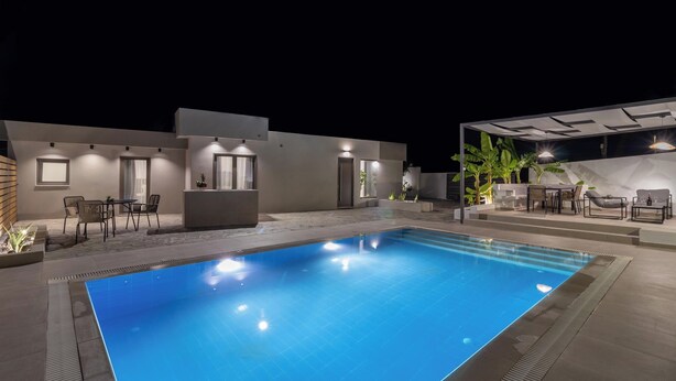 A swimming pool with a tiled deck, a seating area with chairs and a table, and a covered outdoor space with plants and lighting. - Melia Villa in Kolympia (Kolympia)